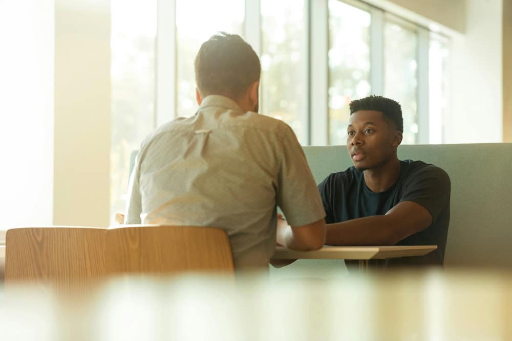 Two men sit across from each other at a table in a brightly lit room, having a conversation. Two men sit across from each other at a table in a brightly lit room, having a conversation.