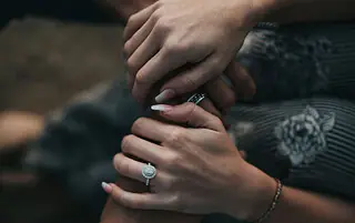 Close-up of three hands gently holding each other, with one hand wearing a diamond engagement ring and another hand wearing a patterned bracelet.