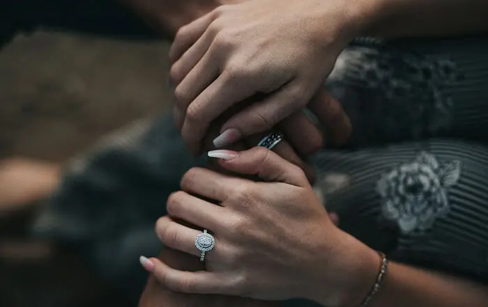 Close-up of three hands gently holding each other, with one hand wearing a diamond engagement ring and another hand wearing a patterned bracelet.